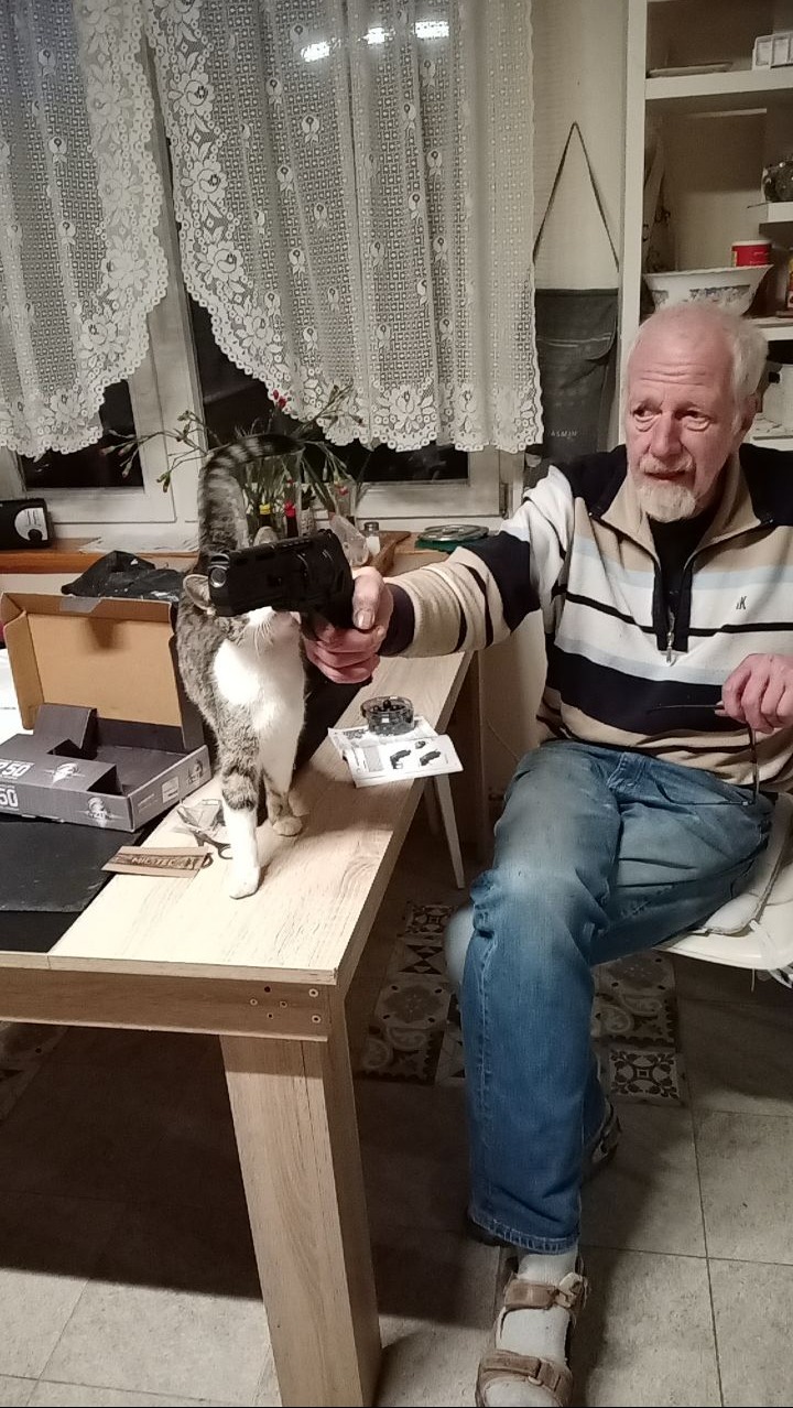 Mężczyzna w swetrze w paski i jeansach trzyma w ręku czarny pistolet, a kot stoi na stole i wącha go; obok leżą instrukcje i akcesoria.