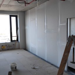 Łukasz Czarnik Mal-Mont Gips - Wnętrze w trakcie prac wykończeniowych w Krakowie: ściany pokryte płytami, widoczne profile metalowe na podłodze, drabina oparta o ścianę, instalacja przeciwpożarowa pod sufitem.