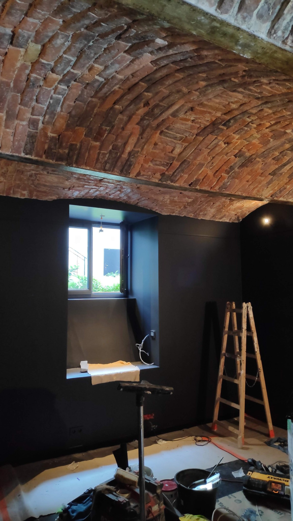 Wnętrze z ceglanym sklepieniem łukowym, pomalowane ściany na ciemny kolor, widoczne narzędzia i drabina, przygotowane do dalszych prac wykończeniowych.
