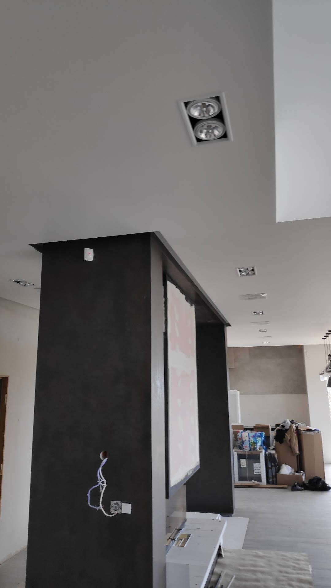 Ciemnoszara kolumna z gładzią, widoczne przewody elektryczne i sufit z oświetleniem punktowym, remont w trakcie.