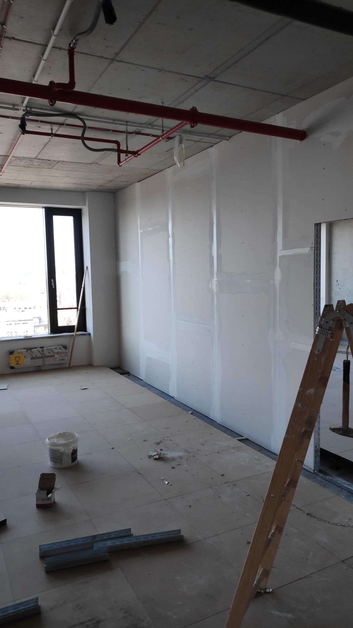 Wnętrze w trakcie prac wykończeniowych w Krakowie: ściany pokryte płytami, widoczne profile metalowe na podłodze, drabina oparta o ścianę, instalacja przeciwpożarowa pod sufitem.