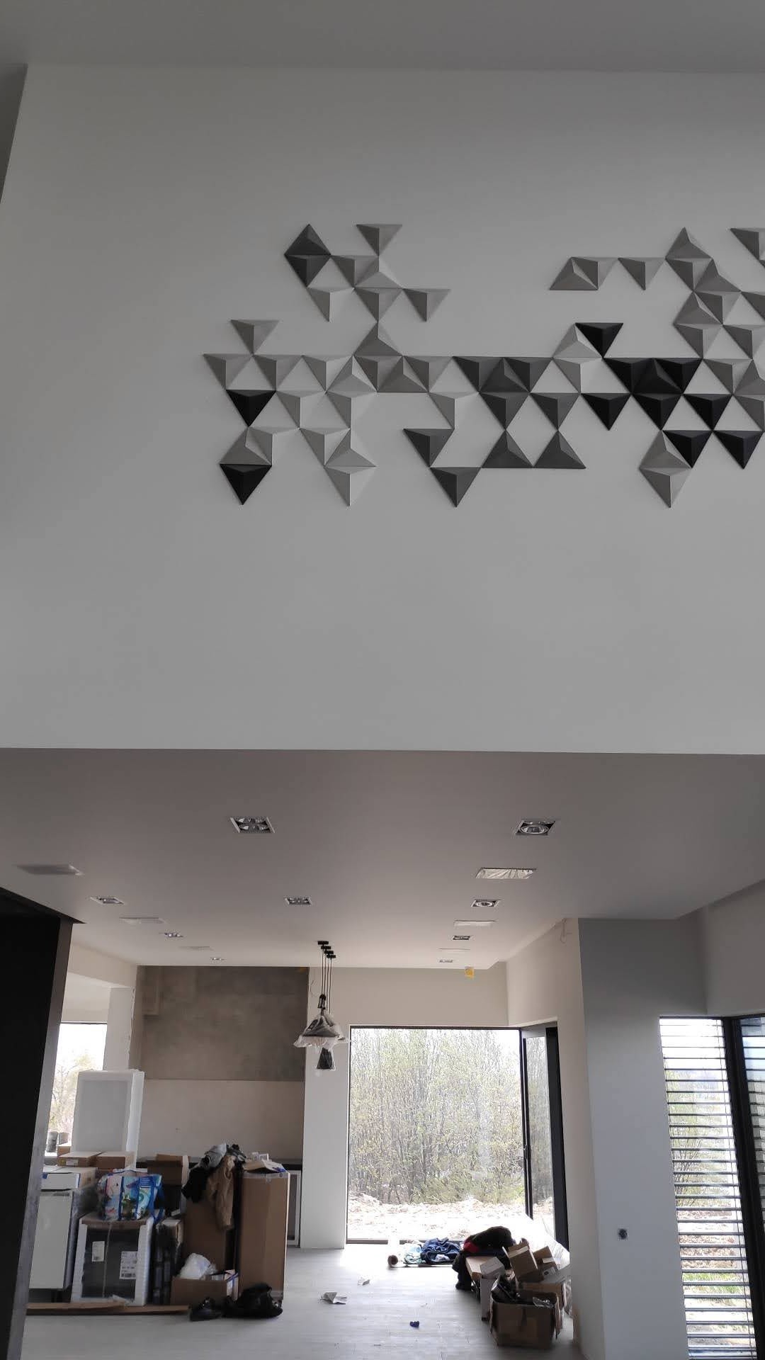 Nowoczesne wnętrze z dekoracją ścienną z trójkątnych paneli w odcieniach szarości i czerni, widok na otwartą przestrzeń z oknem i fragmentem zabudowy kuchennej w trakcie remontu.