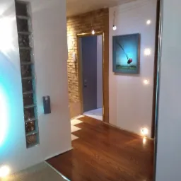 Wnętrze mieszkania z drewnianą podłogą, dekoracyjnymi luksferami w ścianie, obrazem na ścianie i oświetleniem punktowym pod sufitem.