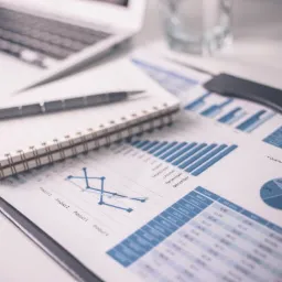 Biurko z laptopem, notatnikiem, długopisem i dokumentami z wykresami słupkowymi, liniowymi i kołowymi prezentującymi dane finansowe.