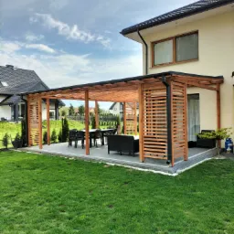 Drewniana pergola z ażurowymi ściankami przy domu jednorodzinnym, z kompletem mebli ogrodowych na szarym brukowanym tarasie, widok na zadbany trawnik i sąsiednie budynki w tle.