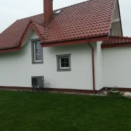 Dom jednorodzinny z białą elewacją, czerwoną dachówką i szarymi ramami okiennymi, na trawniku leży pies rasy mieszanej, widoczna jednostka zewnętrzna klimatyzacji na metalowym stelażu.