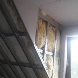 Skośne poddasze w trakcie izolacji: widoczne metalowe profile, wełna mineralna między nimi oraz częściowo zamontowane płyty. W rogu okno z widokiem na zieleń.