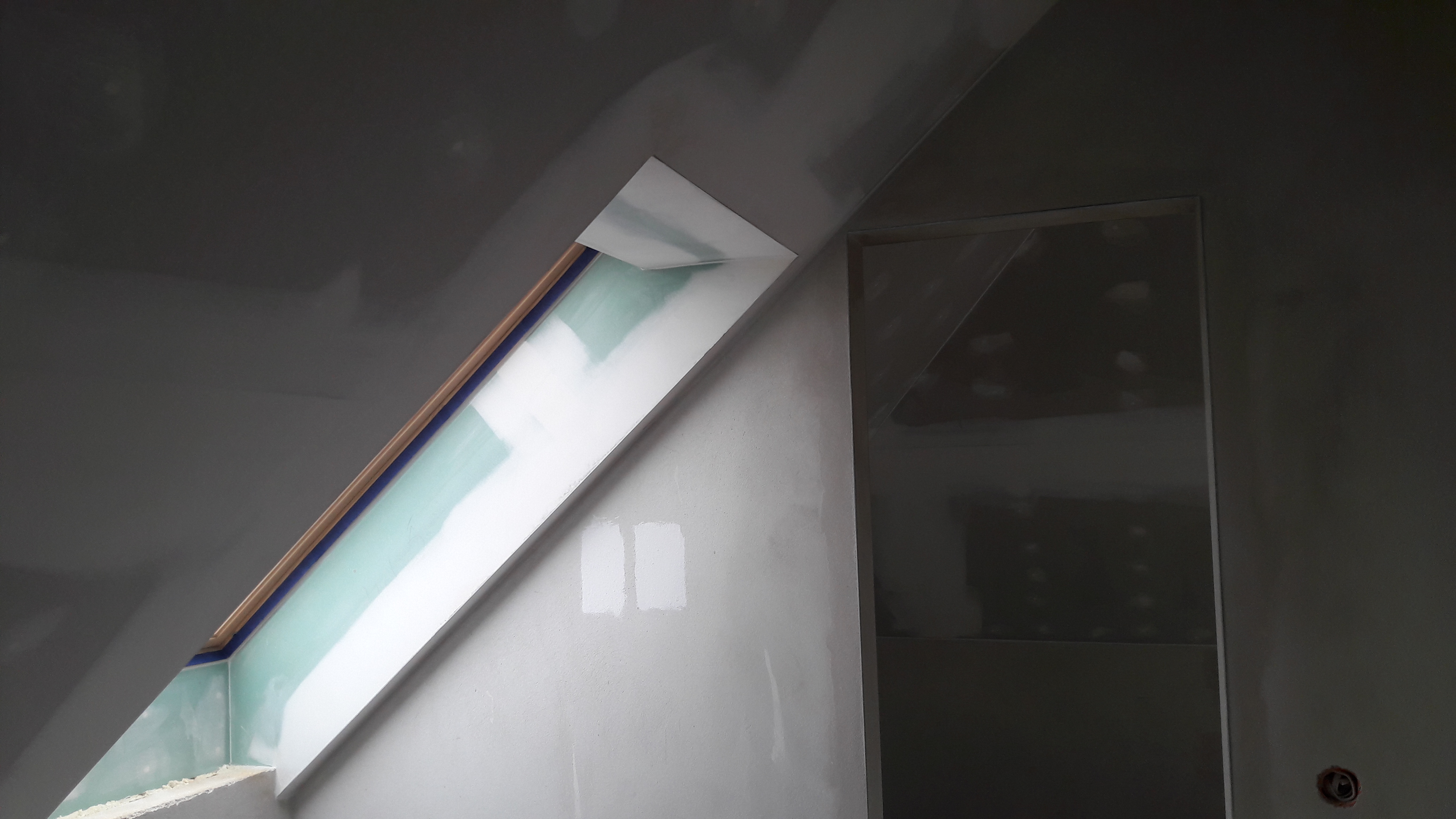 Poddasze w trakcie wykańczania: okno dachowe obudowane płytami, widoczne szpachlowanie i łączenia płyt. Ściany przygotowane do malowania, widoczne próbniki koloru.