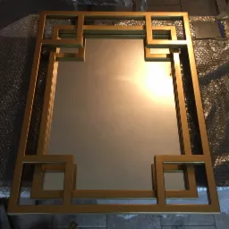 Złota rama lustra z geometrycznym wzorem kwadratów, leżąca na podłożu z folii bąbelkowej i płytek.