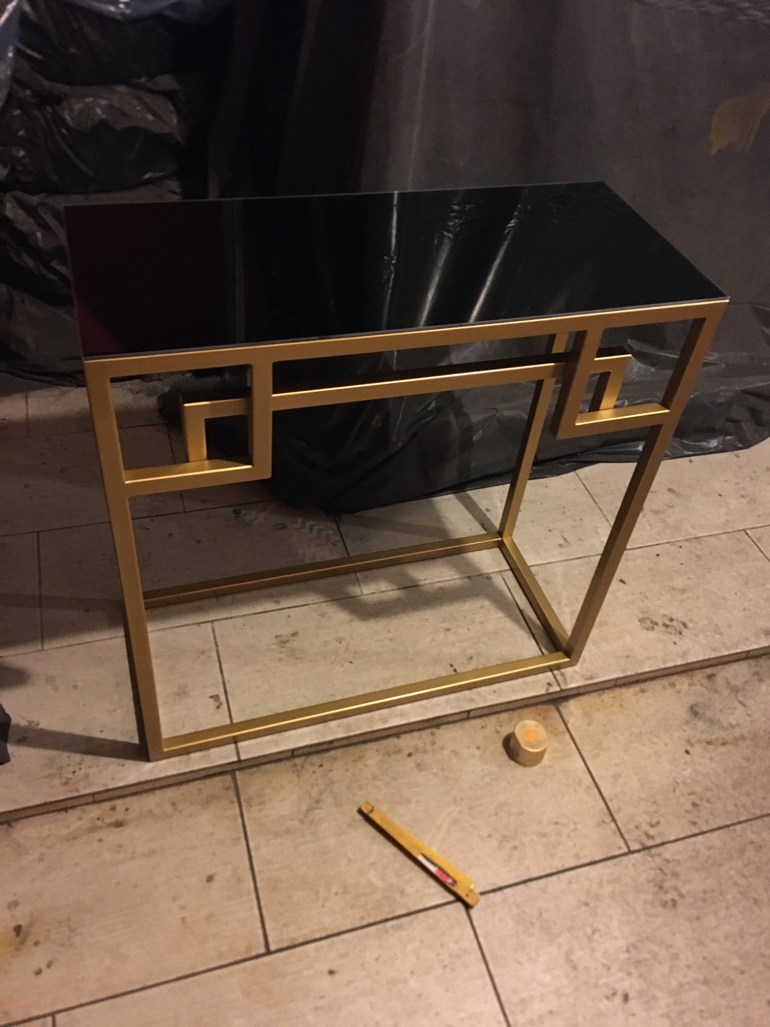 Złoty, metalowy stolik kawowy z czarnym, błyszczącym blatem, o geometrycznym designie, stojący na płytkach podłogowych, z widocznym obok narzędziem pomiarowym i elementem mocującym.