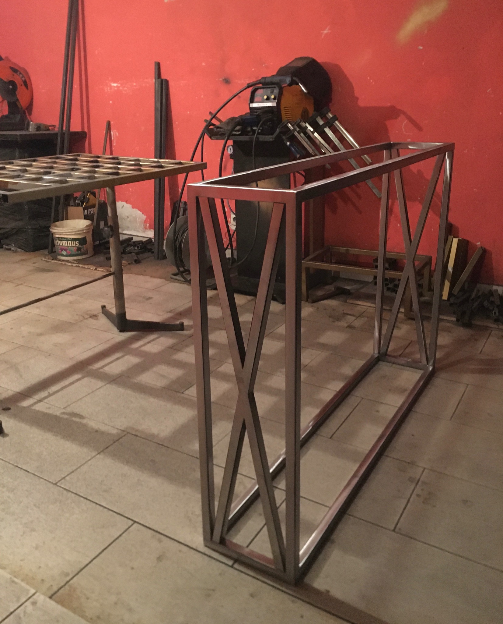 Metalowa konstrukcja stołu z krzyżowymi wzmocnieniami, spawarka i inne narzędzia w tle na tle czerwonej ściany.