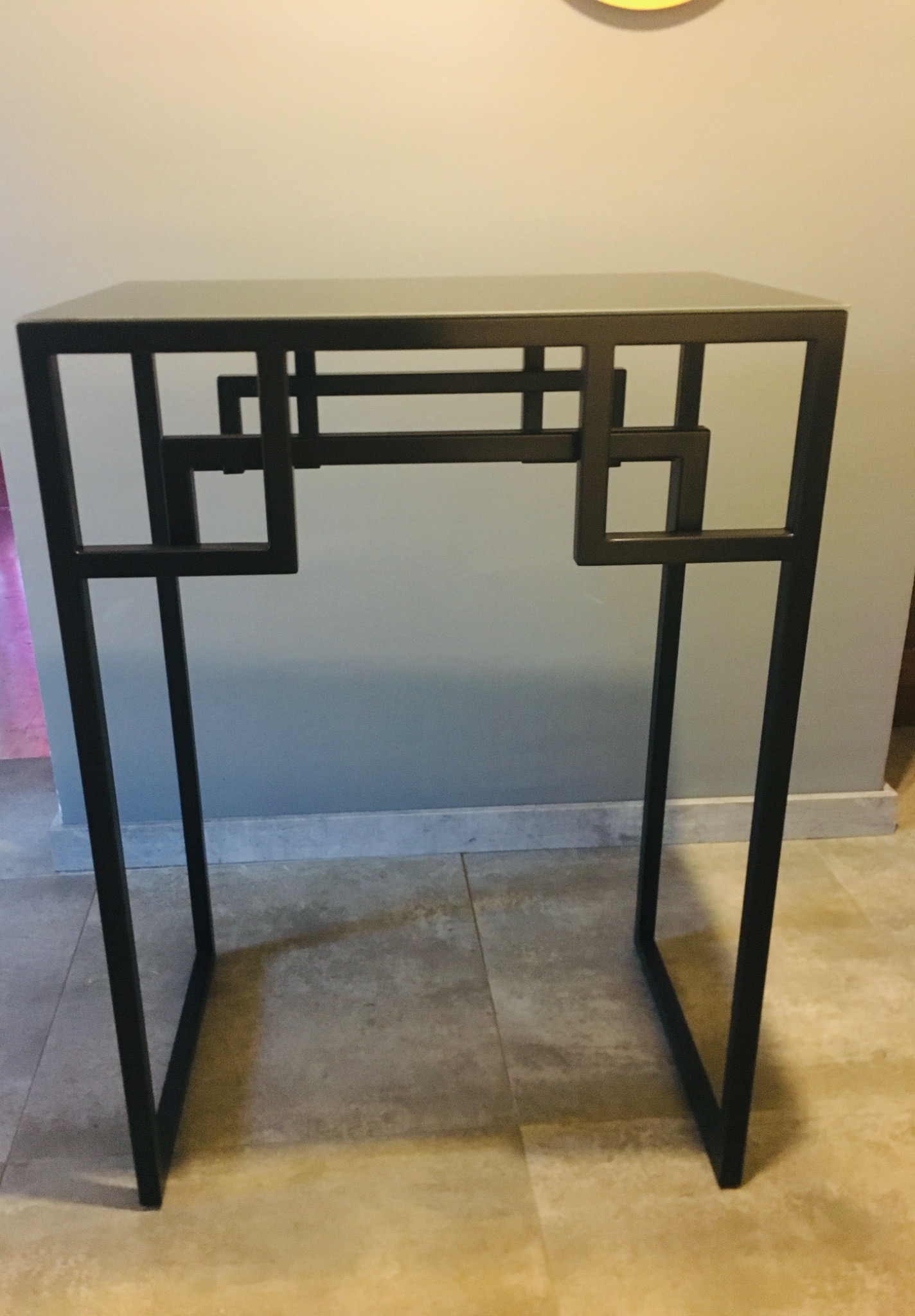 Metalowy stolik pomocniczy o geometrycznym wzorze spawanych profili, czarny mat, na tle beżowej ściany i podłogi.