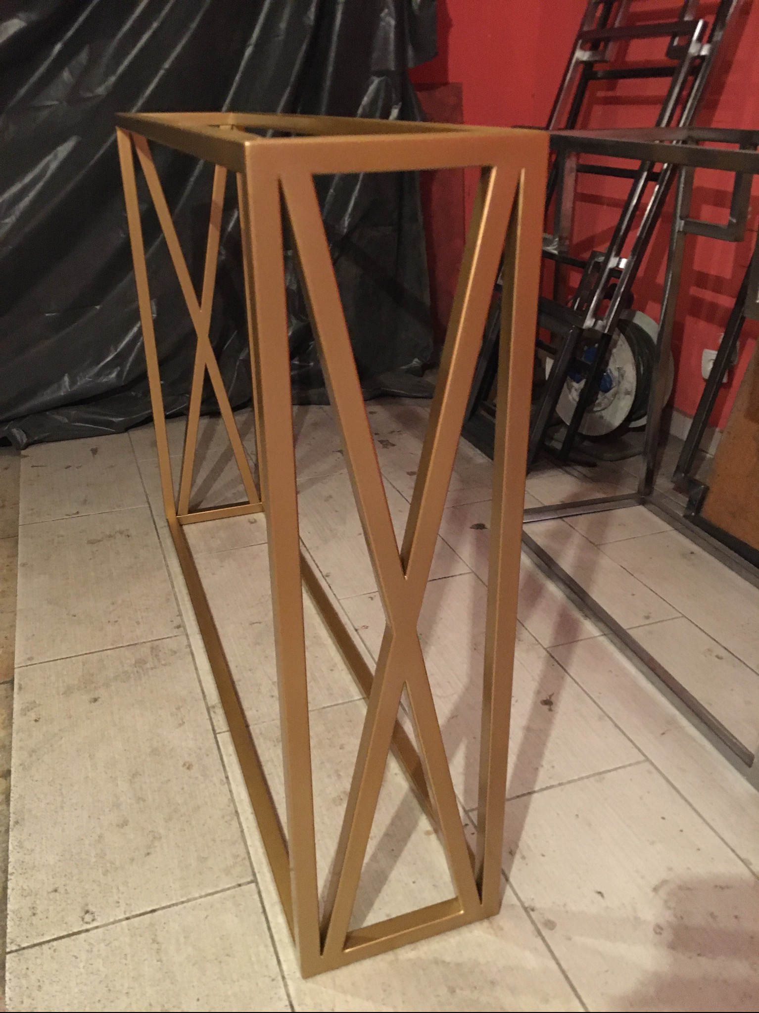 Złota, metalowa konstrukcja stolika z krzyżowymi wzmocnieniami, widoczna w warsztacie na tle czerwonej ściany i innych metalowych elementów.