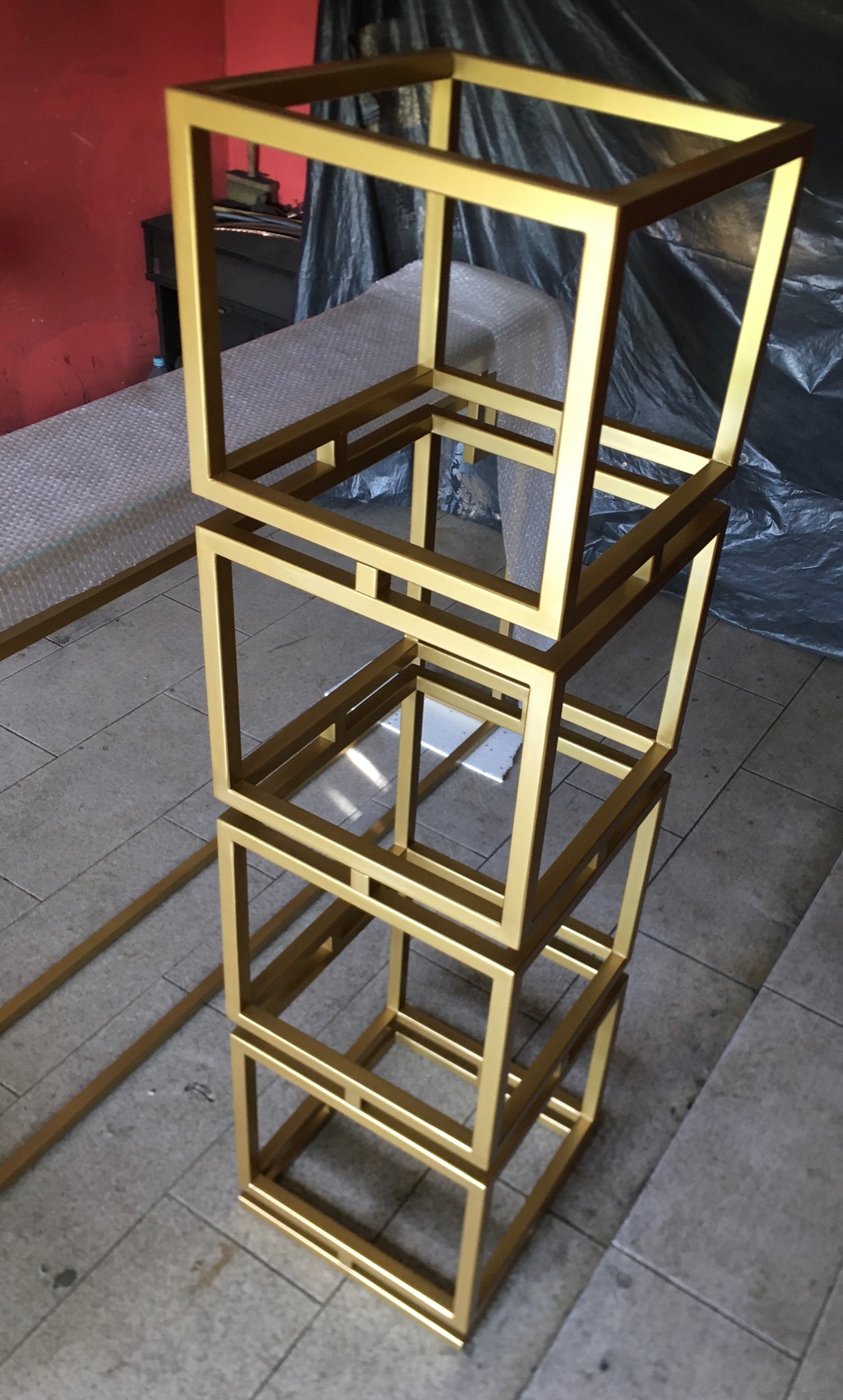 Konstrukcja z trzech złotych, metalowych sześcianów ustawionych jeden na drugim, z dodatkowym elementem leżącym obok na szarej podłodze.