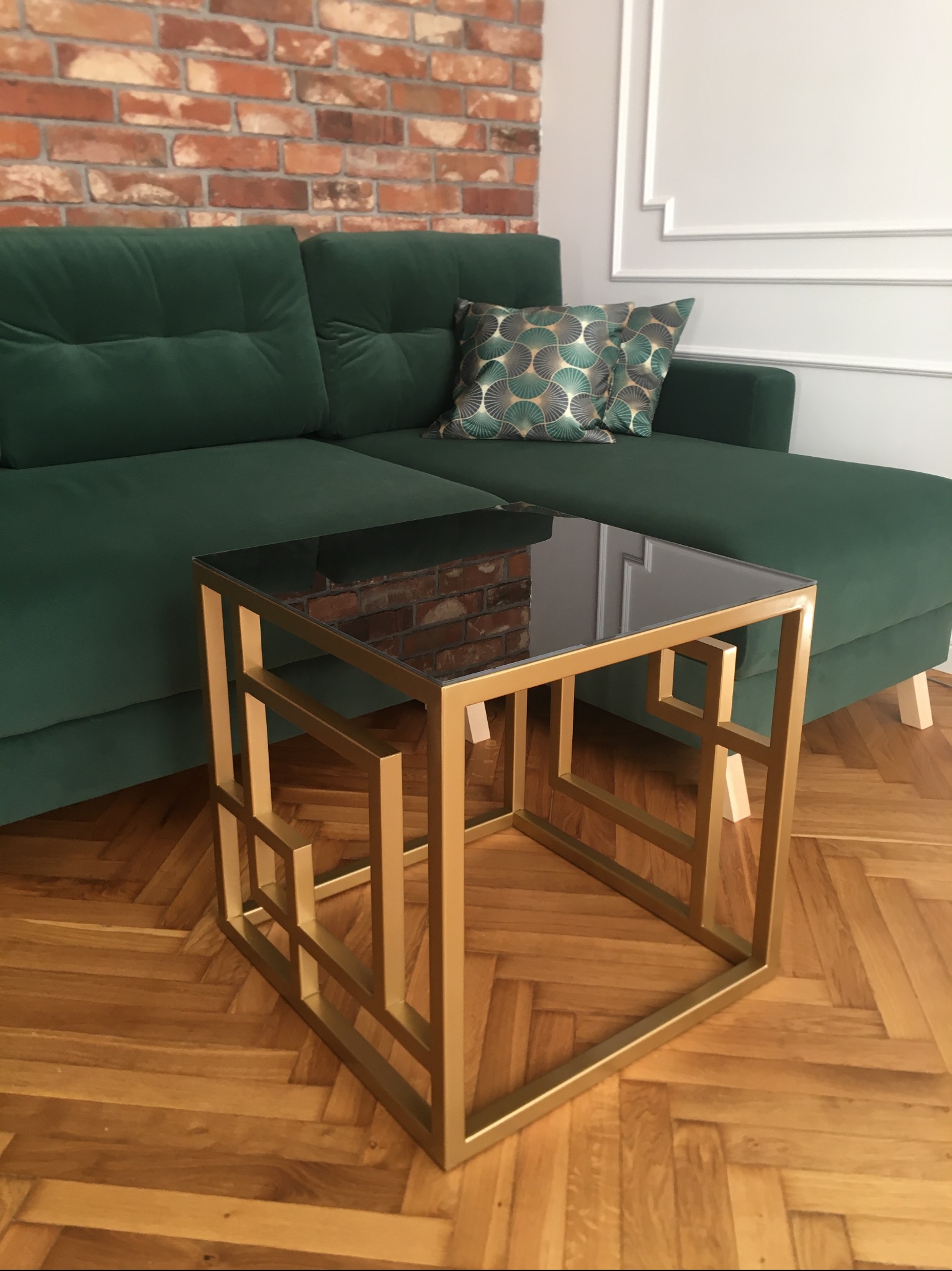 Kwadratowy stolik kawowy ze złotym, geometrycznym stelażem i czarnym, szklanym blatem, ustawiony obok zielonej, welurowej sofy narożnej na parkiecie w jodełkę.