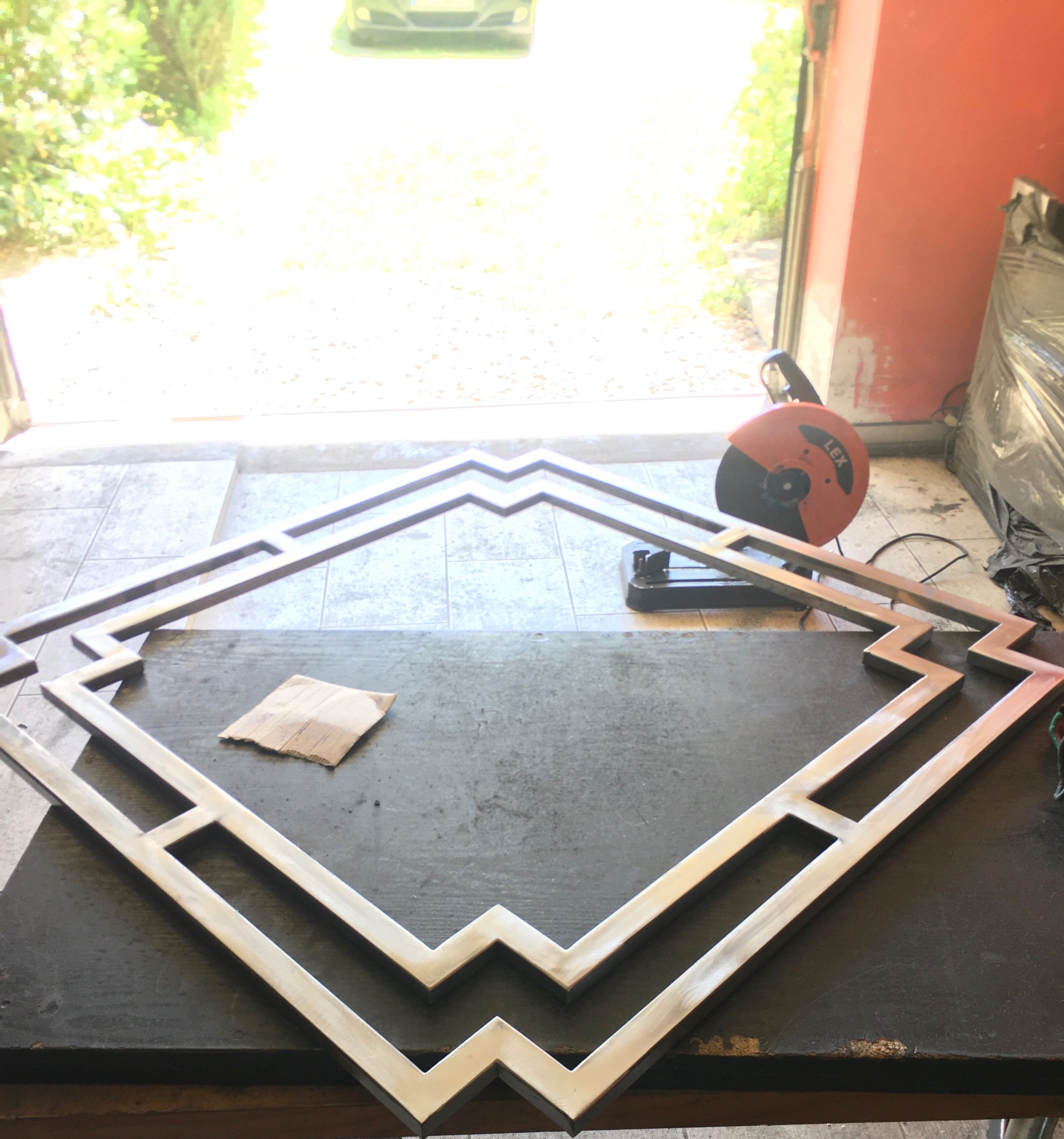 Metalowa konstrukcja o geometrycznym wzorze, prawdopodobnie element dekoracyjny, leżąca na warsztatowym stole, obok widoczna szlifierka kątowa i kawałek papieru ściernego.