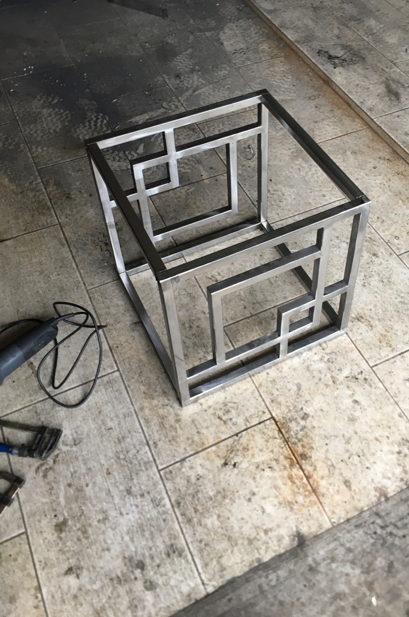 Stalowa, spawana konstrukcja sześcianu z geometrycznym wzorem wewnętrznym, widoczna na podłodze z płytek. Obok leży szlifierka kątowa z przewodem.