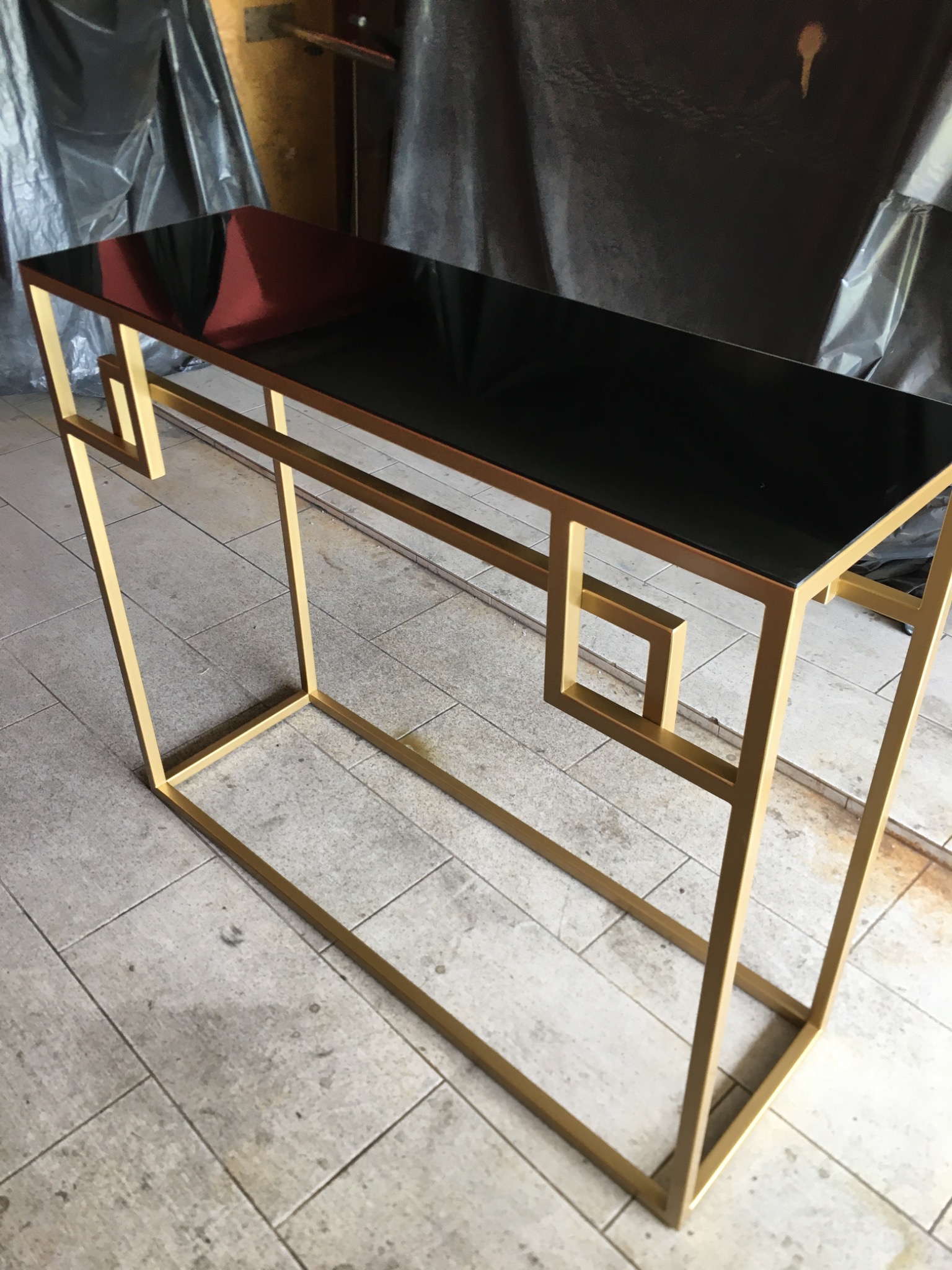 Metalowa konsola z czarnym szklanym blatem, złote profile o geometrycznym kształcie, na tle szarej podłogi z płytek i czarnej folii.