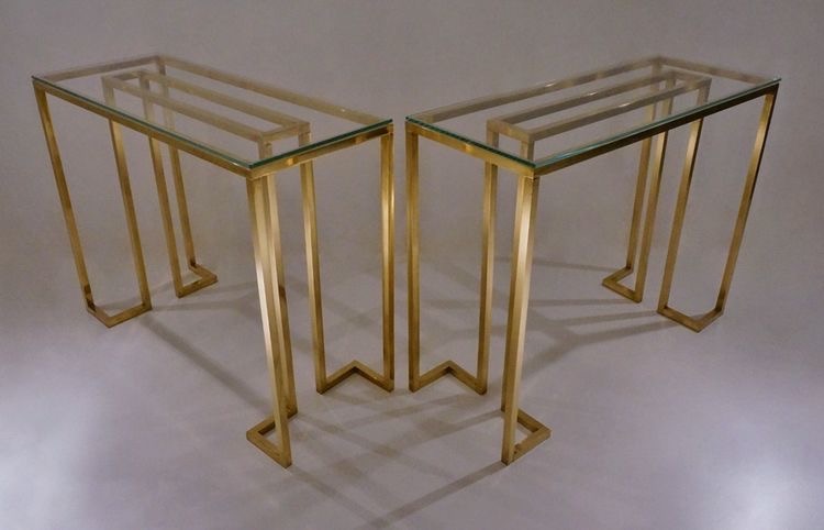 Dwa identyczne stoliki konsolowe z szklanymi blatami i podstawami z polerowanego, złotego metalu o geometrycznym kształcie, ustawione na jasnym tle.