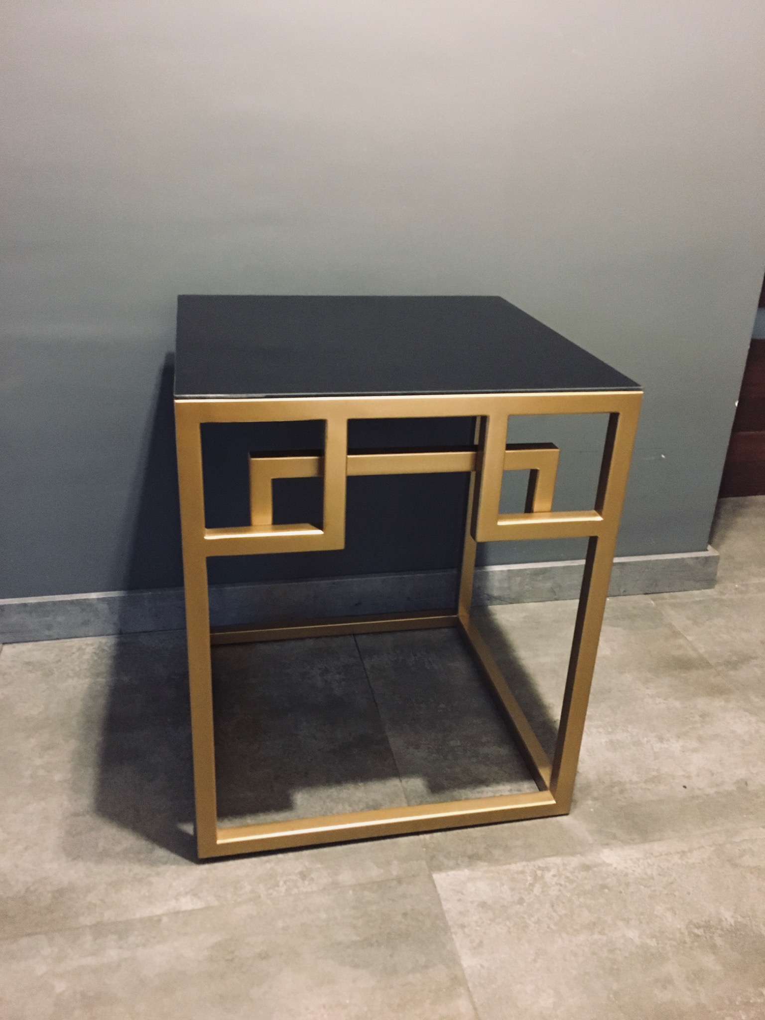 Mały stolik kawowy z czarnym blatem i złotą, geometryczną ramą w stylu art deco, ustawiony na szarej podłodze.