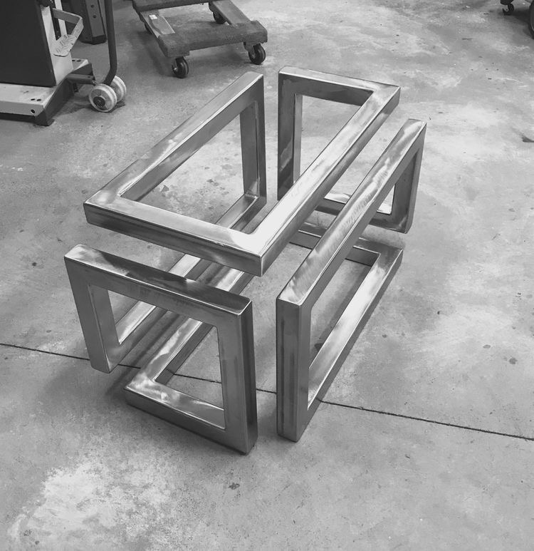 Czarno-białe zdjęcie spawanej, metalowej konstrukcji przestrzennej z profili kwadratowych, przypominającej niedokończony sześcian, leżącej na betonowej podłodze warsztatu, z wózkiem transportowym...