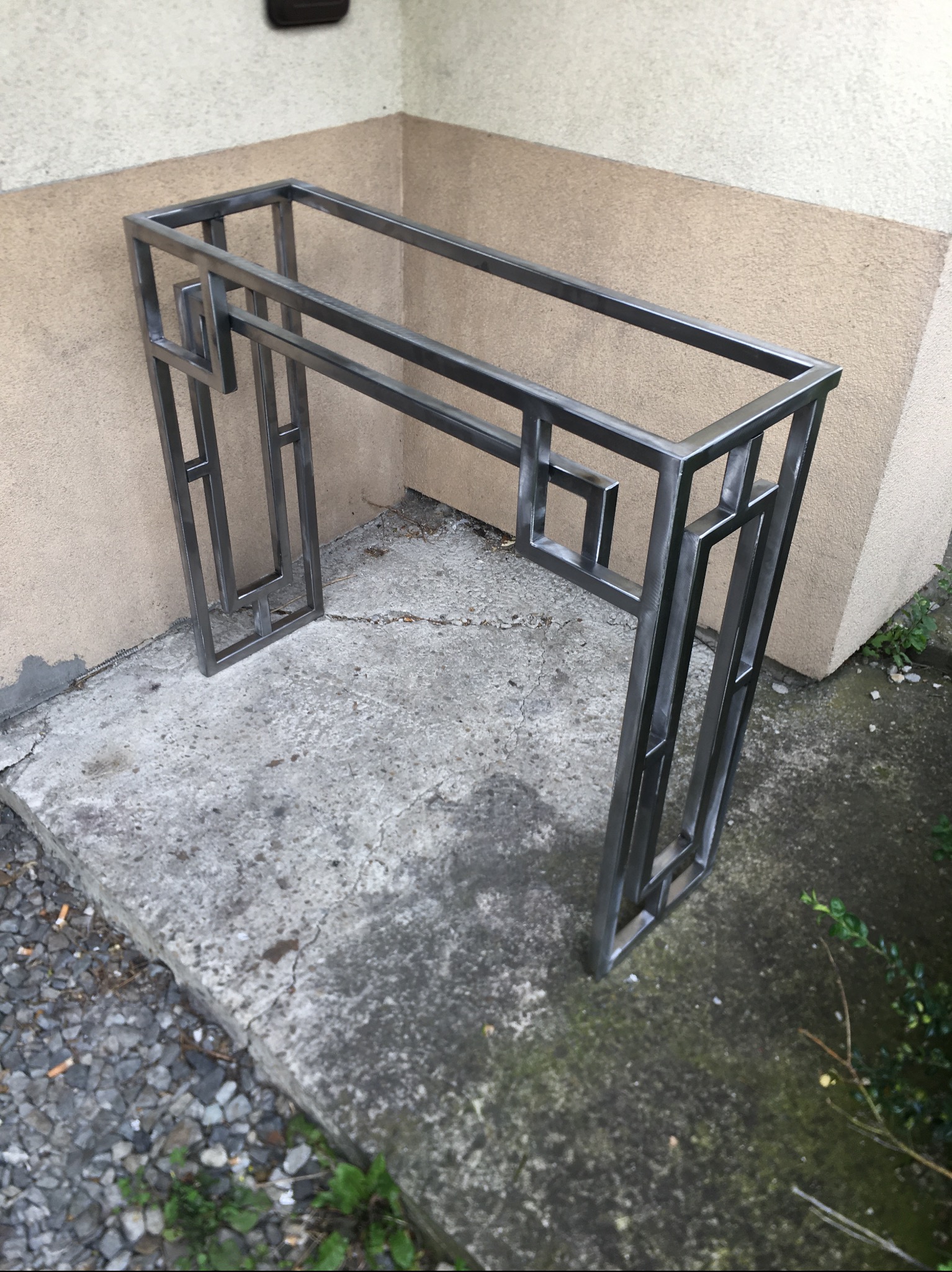 Metalowa konsola o geometrycznym wzorze, wykonana z surowej stali, ustawiona na betonowej posadzce przy ścianie budynku.