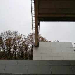 Fragment elewacji budynku z szarej cegły z widocznym betonowym stropem i drzewami w tle, pochmurny dzień.