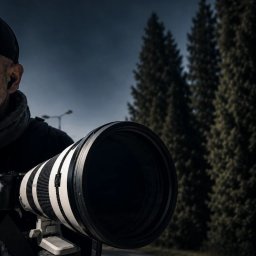 Biuro Detektywistyczne PS - Osoba w czapce z daszkiem trzymająca duży, biało-czarny teleobiektyw na statywie, skierowany w dal. W tle ciemne drzewa i zachmurzone niebo, sugerujące obserwację lub monitoring.