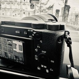 Biuro Detektywistyczne PS - Czarno-białe ujęcie tyłu aparatu bezlusterkowego Sony, którego ekran wyświetla fasadę budynku z balkonami, na tle rozmytego miejskiego krajobrazu z ulicznym znakiem.