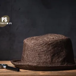 Brązowy kapelusz fedora i lupa leżące na drewnianym stole na tle ciemnej ściany z logotypem w górnym rogu.