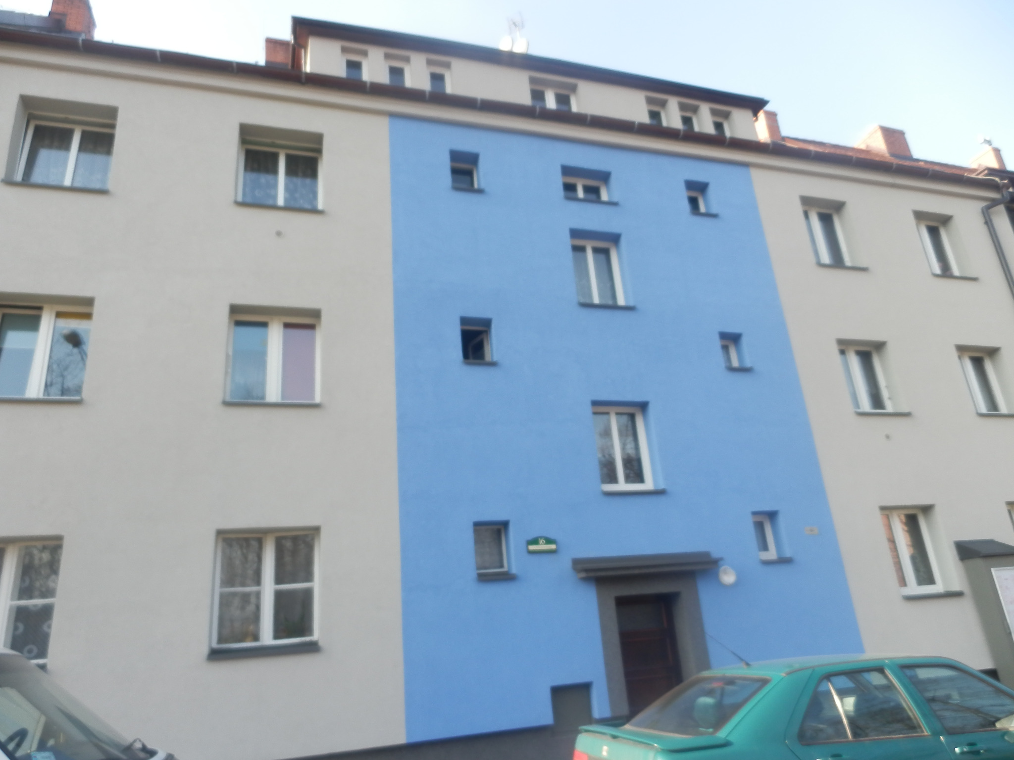 Odnowiona fasada budynku mieszkalnego z wyróżniającym się, niebieskim pasem pośrodku. Widoczne okna i drzwi wejściowe. Częściowo zasłonięty samochód na pierwszym planie.