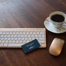 Biurko z drewnianym blatem, na którym leżą: złota klawiatura, myszka, wizytówka z napisem HRbit oraz filiżanka kawy w porcelanowej filiżance z motywem kwiatowym.