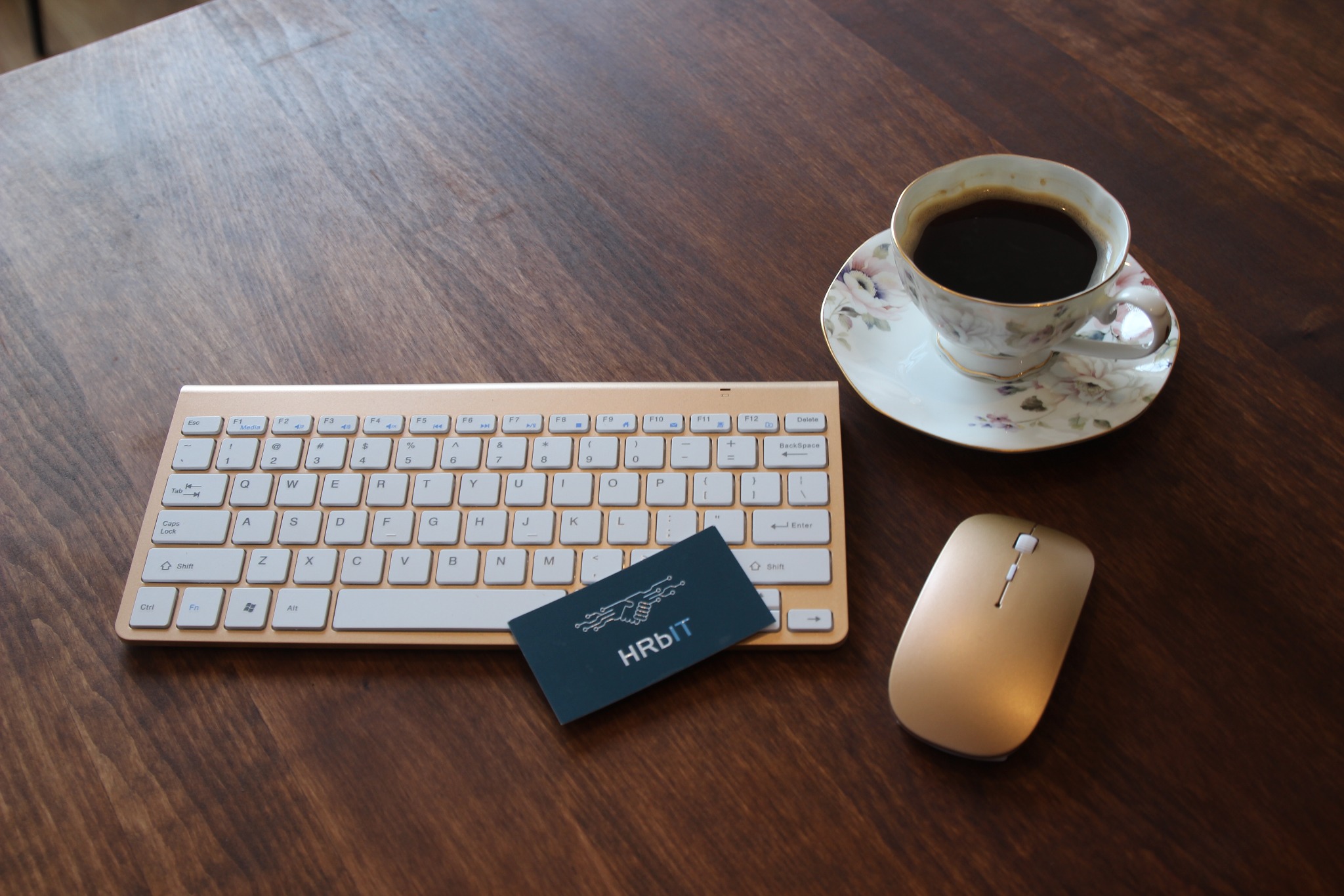 Biurko z drewnianym blatem, na którym leżą: złota klawiatura, myszka, wizytówka z napisem HRbit oraz filiżanka kawy w porcelanowej filiżance z motywem kwiatowym.