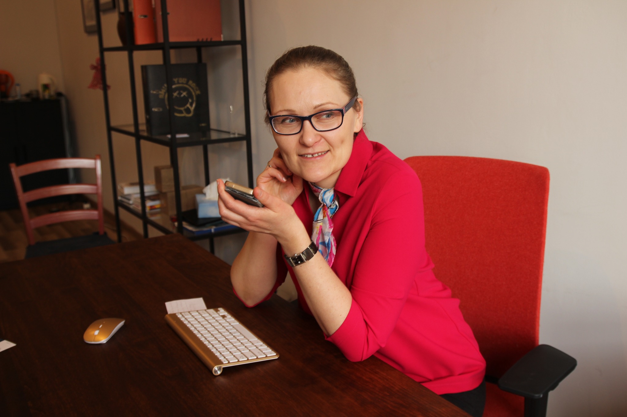 Kobieta w różowej koszuli i okularach siedzi przy biurku w Łodzi, trzymając telefon komórkowy. Na biurku widoczna klawiatura i myszka w kolorze złotym.