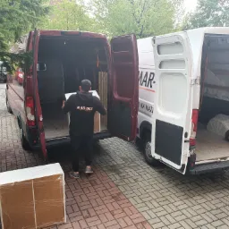 Pracownik w czarnej bluzie z napisem MAR-BUS wnosi zapakowany karton do otwartego bagażnika czerwonego busa, obok stoi biały bus z otwartym bagażnikiem, widoczny wózek transportowy.