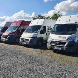 Szereg czterech samochodów dostawczych marki Fiat Ducato firmy Mar-Bus Race zaparkowanych na żwirowym parkingu w słoneczny dzień.