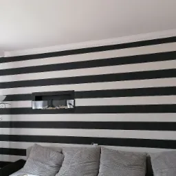 Ściana w czarno-białe poziome pasy z wnęką dekoracyjną, widoczny fragment sofy i lampy stojącej, dekoracyjny gzyms pod sufitem.