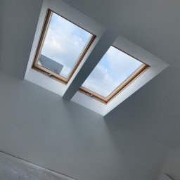 Bartłomiej Pilch - Poddasze z dwoma oknami dachowymi, świeżo pomalowane na szaro, z widocznym fragmentem niezabezpieczonej żarówki i fragmentem podłogi przed wykończeniem.