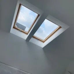 Poddasze z dwoma oknami dachowymi, świeżo pomalowane na szaro, z widocznym fragmentem niezabezpieczonej żarówki i fragmentem podłogi przed wykończeniem.