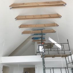 Bartłomiej Pilch - Wnętrze w trakcie malowania z drewnianymi belkami pod sufitem, zabezpieczonymi folią malarską, widoczne rusztowanie i fragment pomalowanej ściany w jasnym odcieniu.