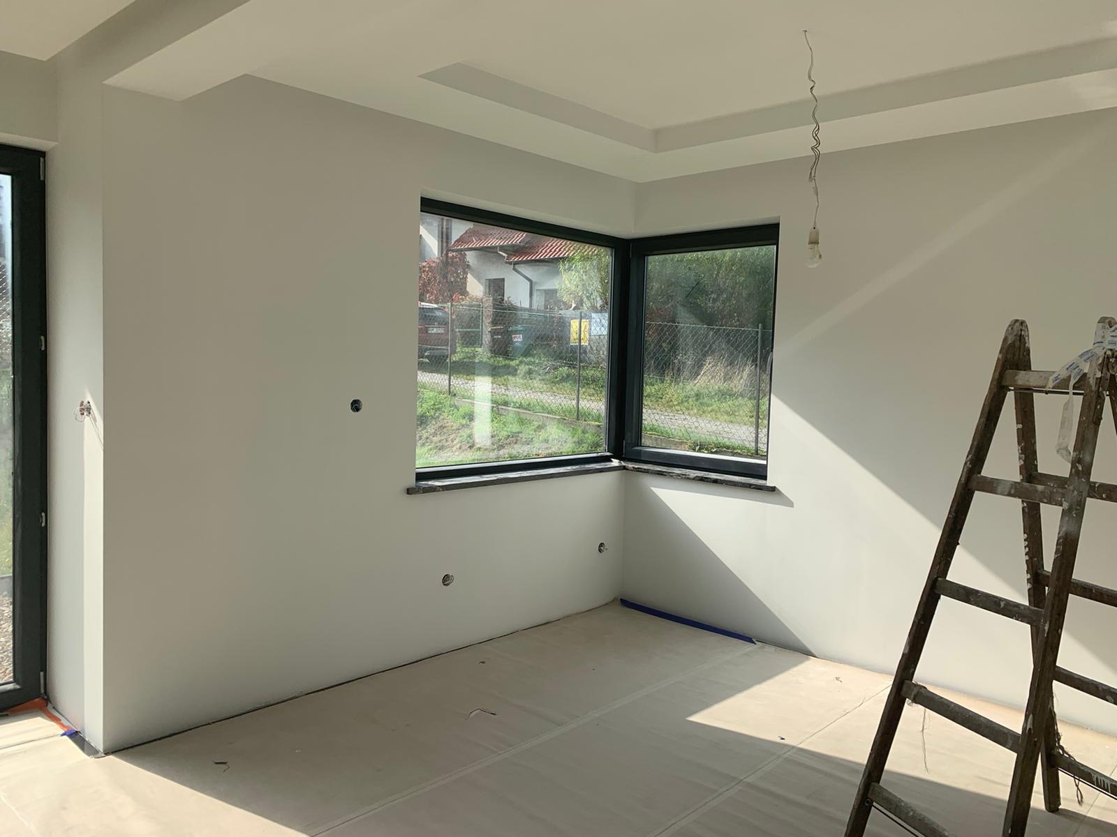 Świeżo pomalowane na biało wnętrze z dużym oknem narożnym, zabezpieczona podłoga, drewniana drabina oparta o ścianę, widoczne puszki instalacyjne.