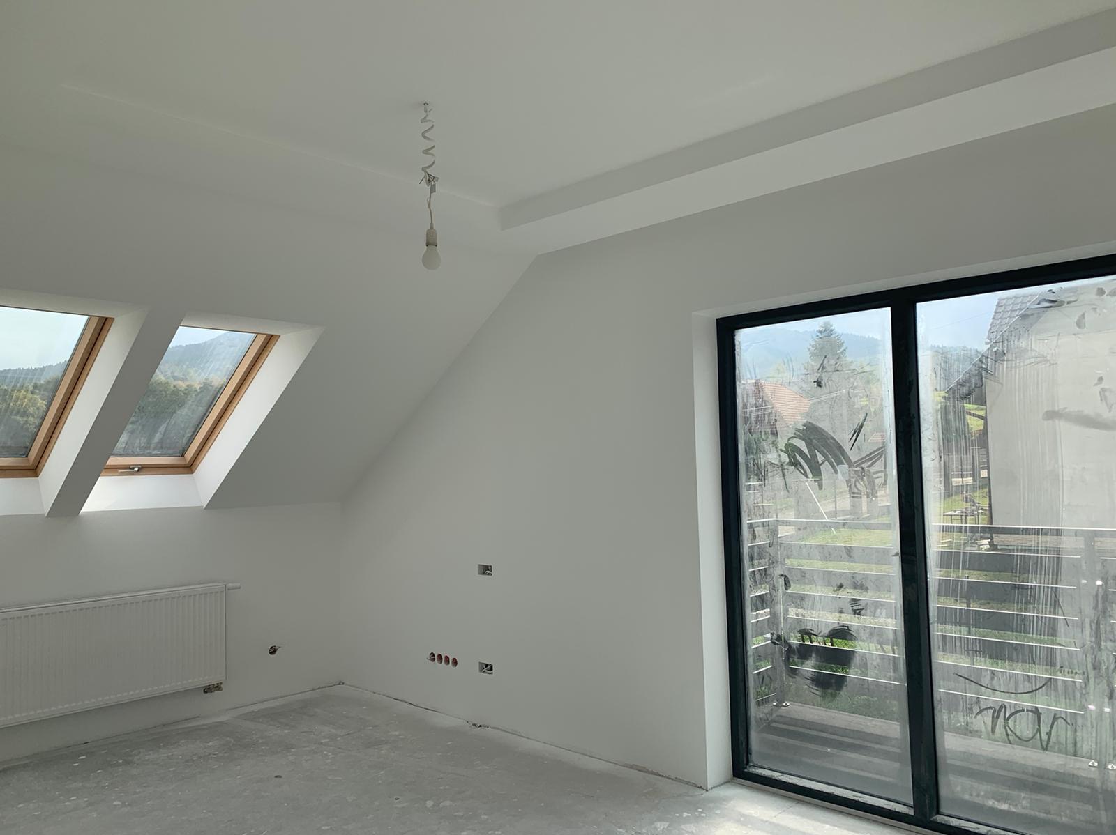Poddasze po świeżym pomalowaniu na biało, widoczne dwa okna dachowe, grzejnik, okno balkonowe z folią zabezpieczającą, surowy beton na podłodze i wisząca żarówka na kablu.