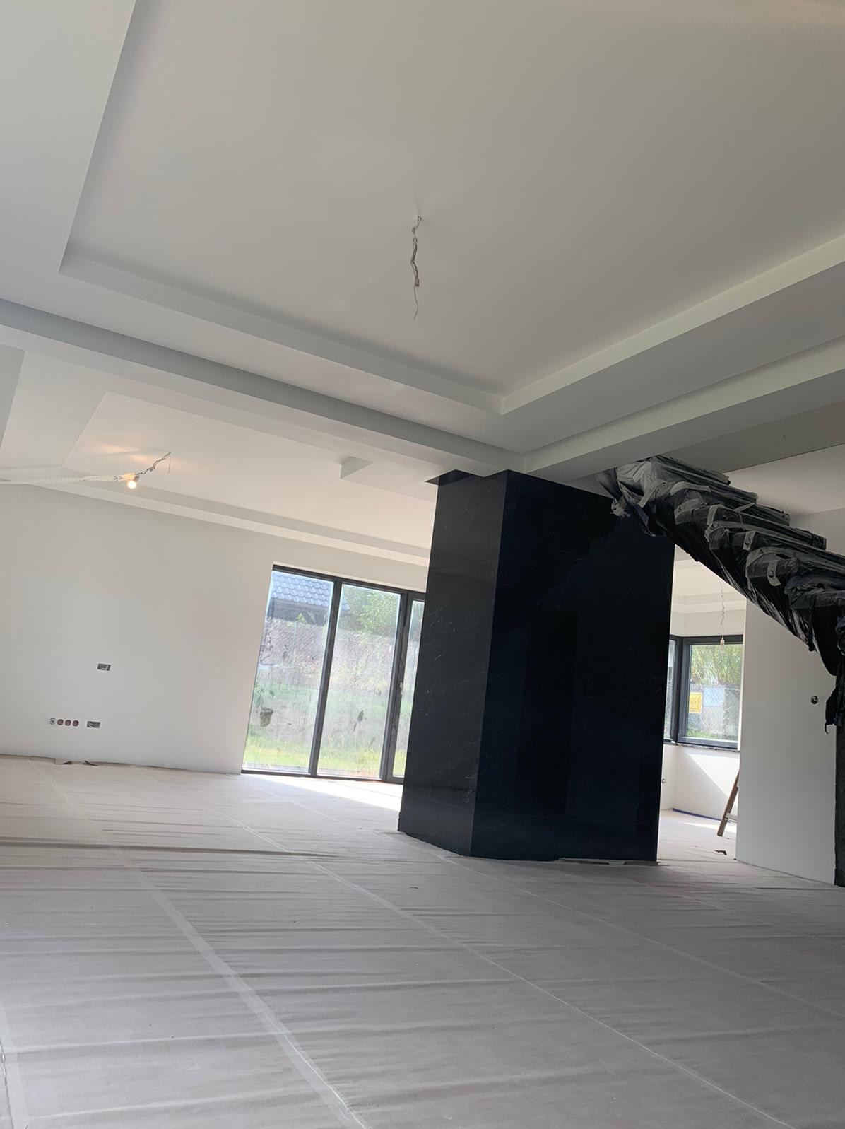 Wnętrze pomieszczenia w trakcie prac wykończeniowych: białe ściany i sufit z dekoracyjnymi listwami, podłoga zabezpieczona folią malarską, czarny filar częściowo osłonięty czarną folią ochronną...