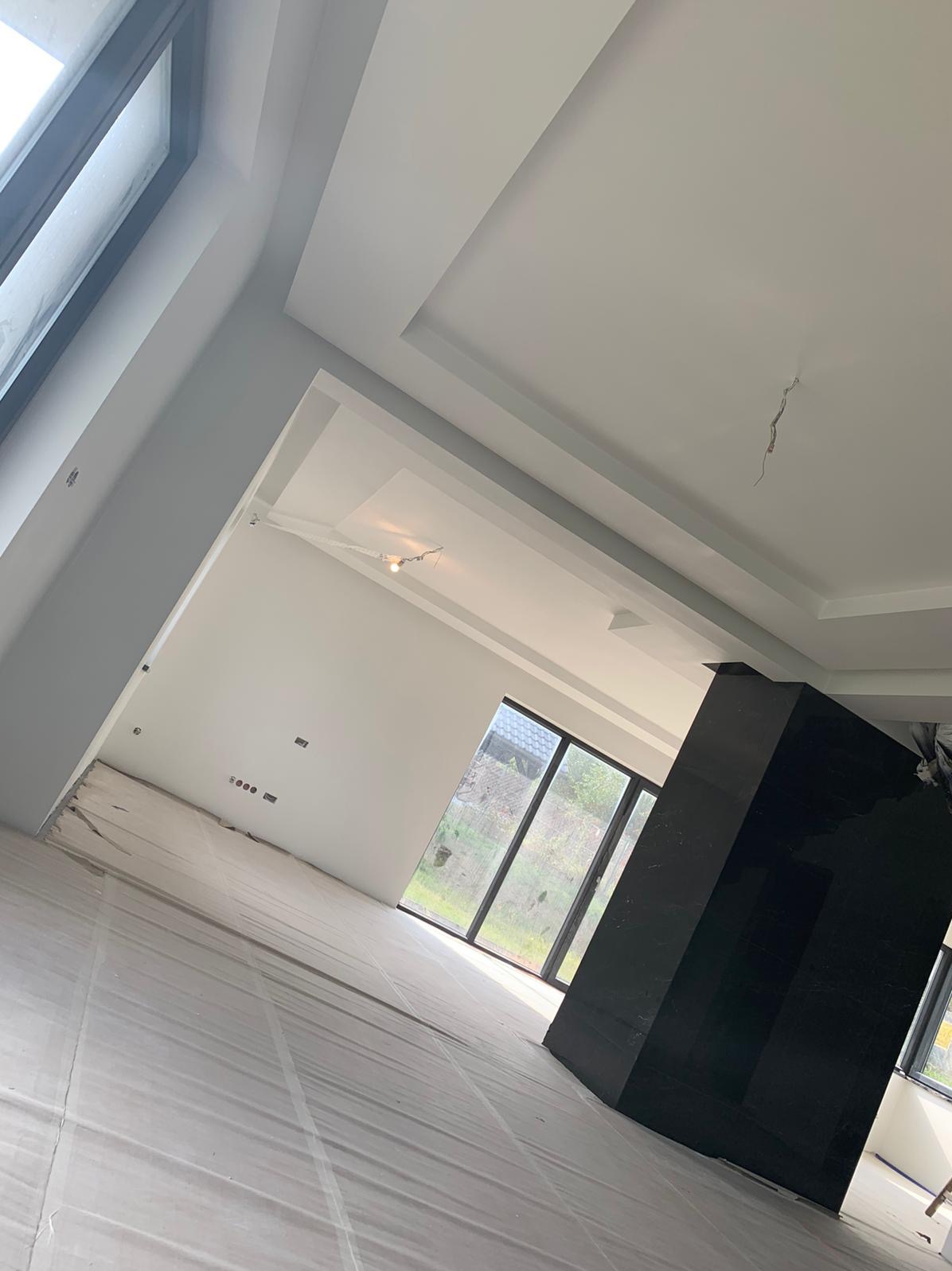 Wnętrze w trakcie malowania, podłoga zabezpieczona folią, ściany świeżo pomalowane na biało, widoczne okna i czarny element dekoracyjny, sufit z podwieszanymi elementami.