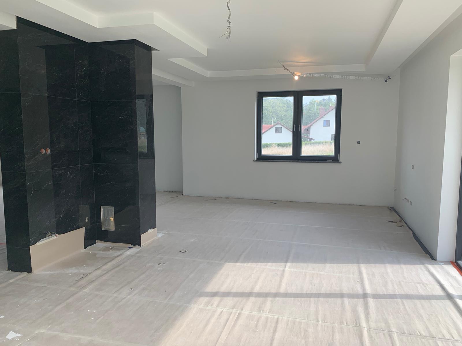 Wnętrze pokoju w trakcie remontu: ściany pomalowane na biało, podłoga zabezpieczona folią ochronną, widoczny fragment kolumny obłożonej czarnym marmurem z wnęką na biokominek.