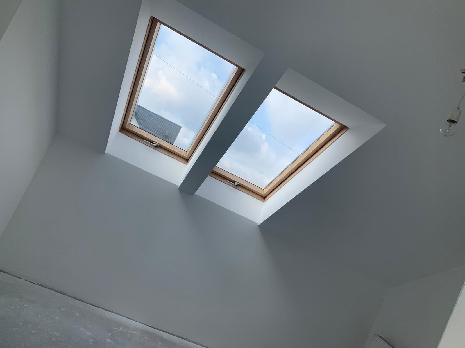 Poddasze z dwoma oknami dachowymi, świeżo pomalowane na szaro, z widocznym fragmentem niezabezpieczonej żarówki i fragmentem podłogi przed wykończeniem.