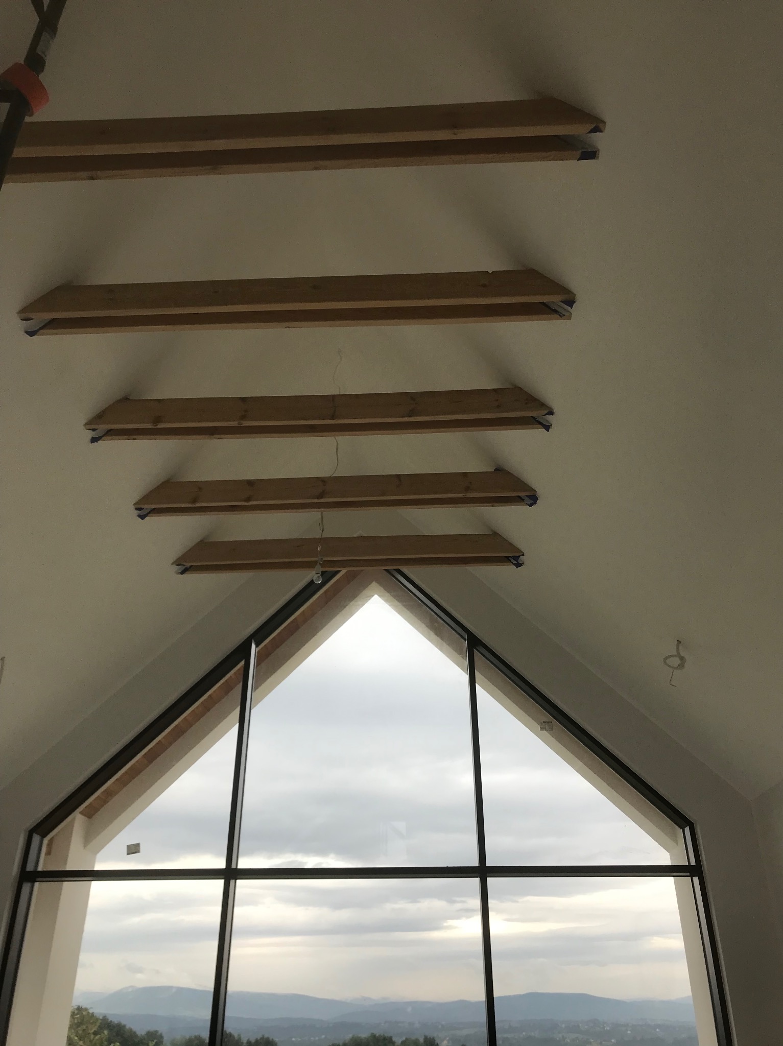 Wnętrze poddasza z drewnianymi belkami zamontowanymi do sufitu, widok na okno z widokiem na krajobraz górski w tle.