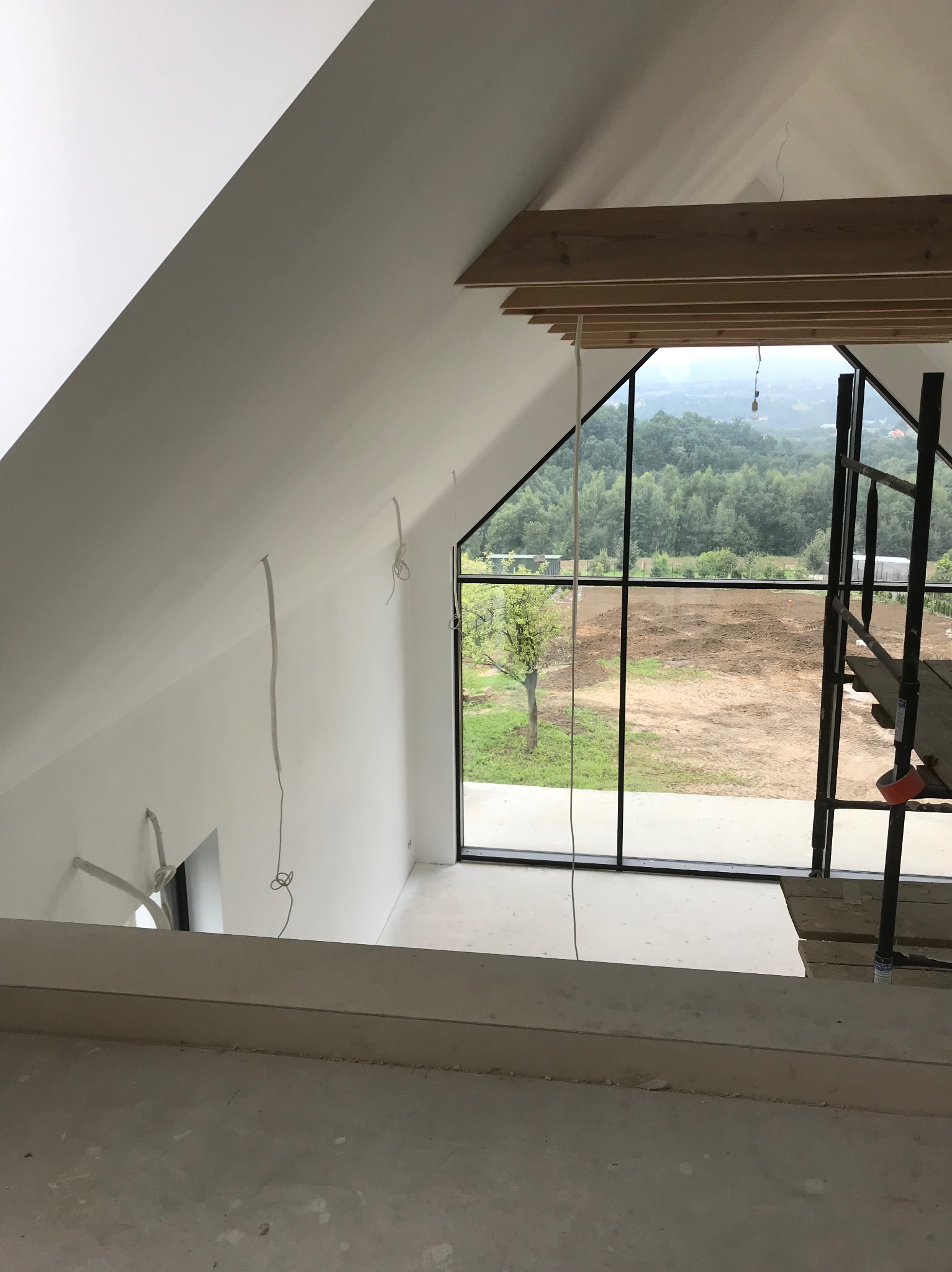 Wnętrze poddasza z dużym oknem i rusztowaniem, ściany przygotowane do malowania, widoczne przewody elektryczne.