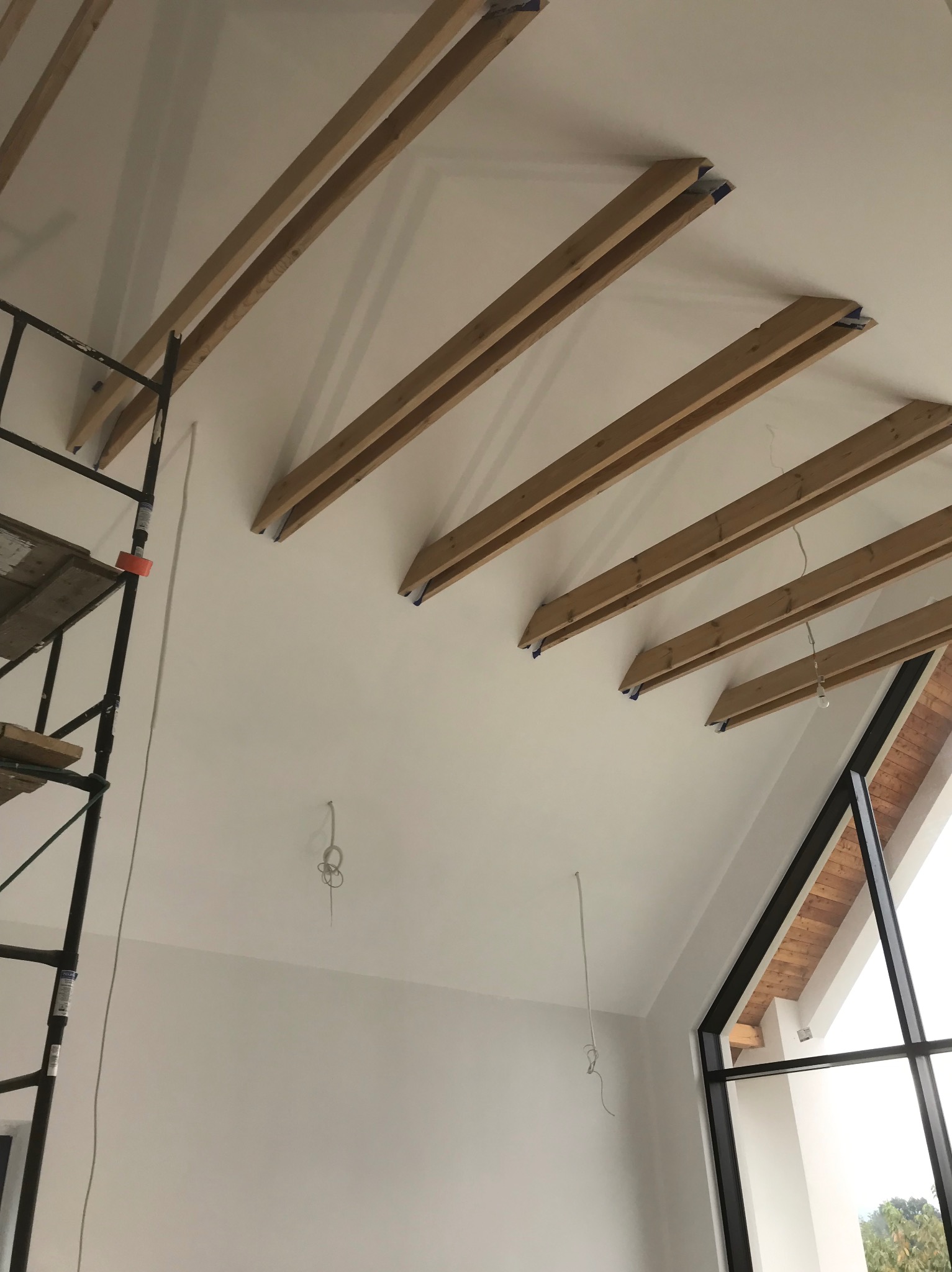 Wnętrze w trakcie prac wykończeniowych: drewniane belki stropowe przymocowane do pomalowanego na biało sufitu, widoczne rusztowanie i przewody elektryczne zwisające z sufitu, duże okno z czarną ramą.