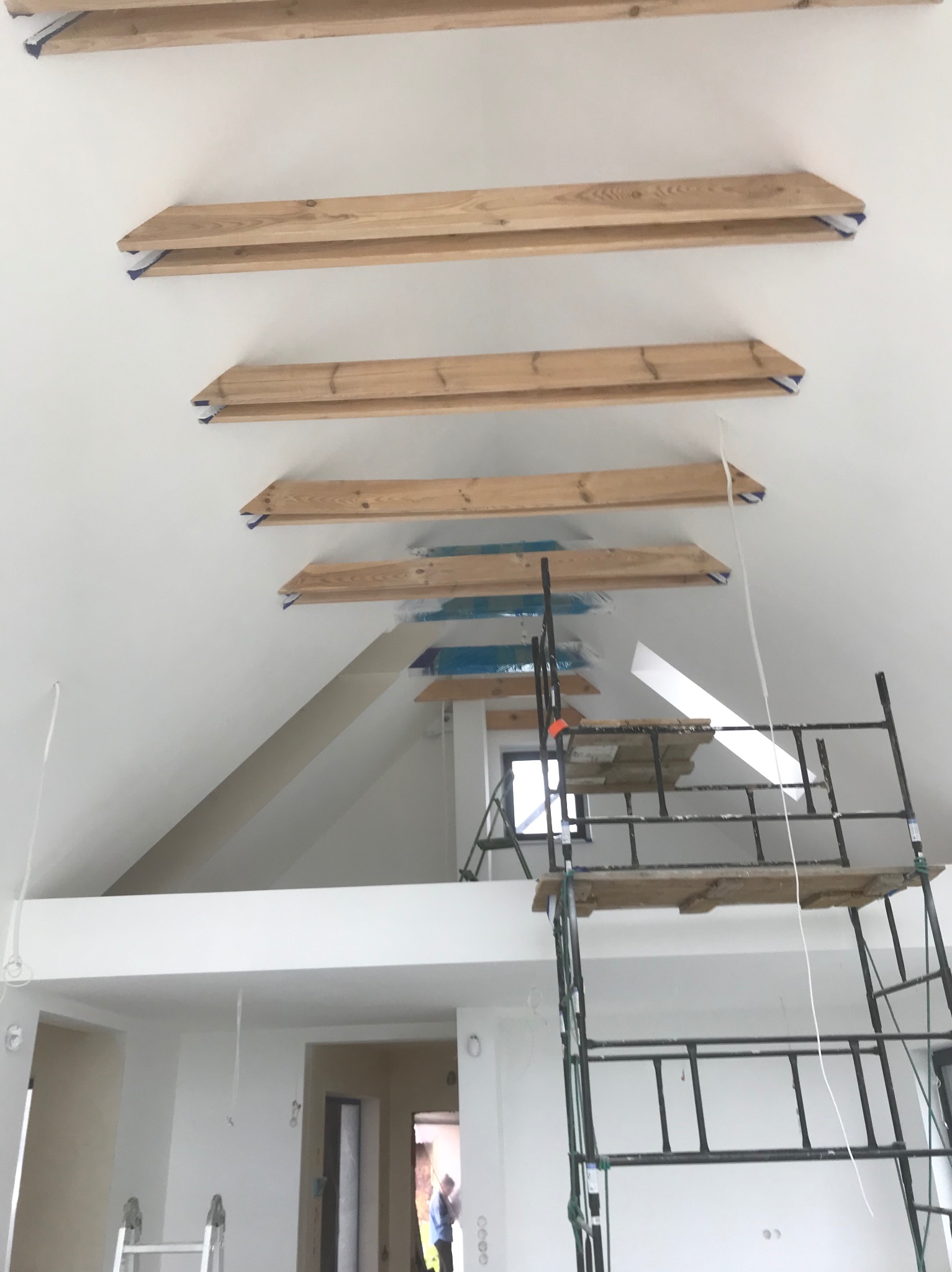 Wnętrze w trakcie malowania z drewnianymi belkami pod sufitem, zabezpieczonymi folią malarską, widoczne rusztowanie i fragment pomalowanej ściany w jasnym odcieniu.